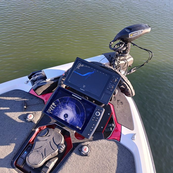 Two fishing electronics screens rigged at the bow of a bass boat, with a trolling motor foot pedal on the deck and the trolling motor head beside them, over open water